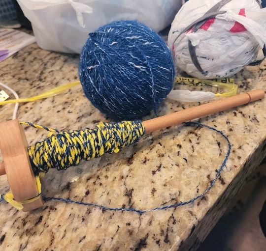 a top whorl drop spindle full of dark blue commercial yarn plied with yellow plastic yarn AKA plarn